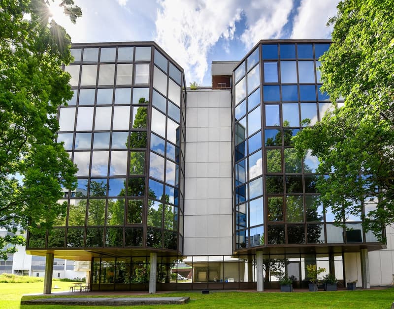 Modern glass and concrete building with large windows, surrounded by green trees and grass. The building has a balcony or terrace visible on the upper floor.
