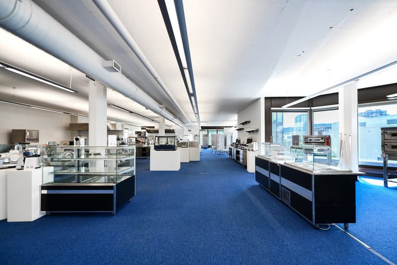 Large open space with blue carpeted floor, various kitchen and home appliances on display, including dishwashers, ovens, and refrigerators. The space has high ceilings with exposed ductwork and large windows providing natural light.