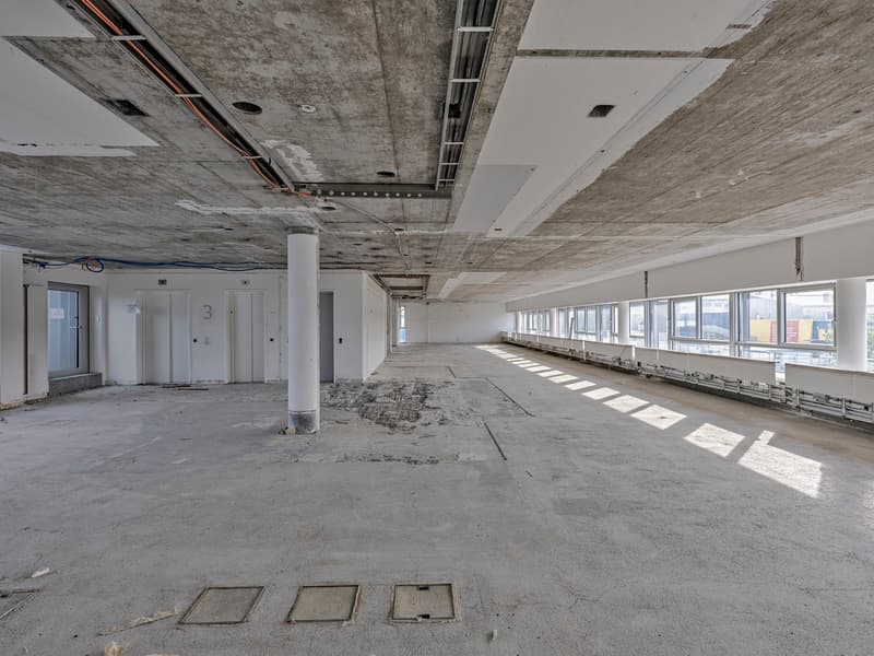 The image shows a large, open commercial space under construction. The space has a concrete floor, exposed concrete ceiling, and several doorways leading to other rooms. The space is well-lit with large windows along one side, providing natural light. The