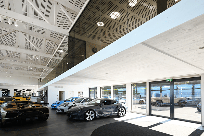 Garage showroom with multiple cars, elevated walkway, glass walls and ceiling, illuminated lights.