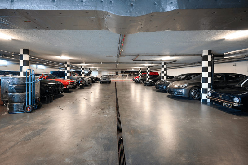 An indoor garage with cars parked, columns, tires on a cart, and overhead lights.