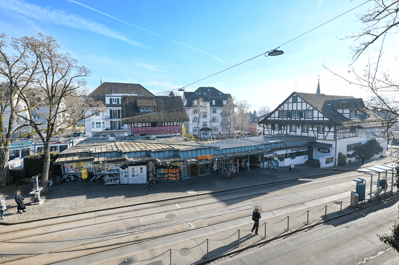 an urban street view with a tram stop and several buildings with shops in the background, large trees and people walking around