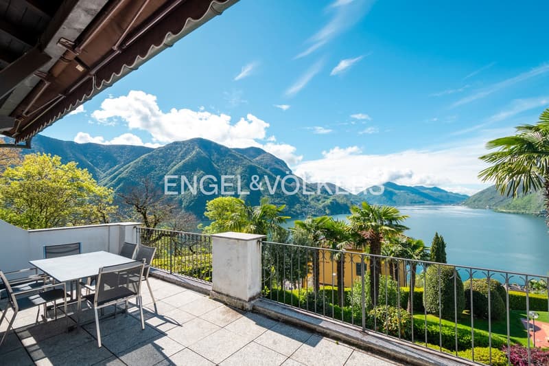 Outdoor terrace with a stunning view of the mountains, lake, and lush greenery. The terrace has a dining table and chairs, and is surrounded by palm trees and other tropical plants.
