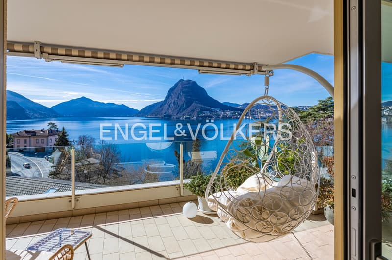 Balcony with large glass windows, view of mountains and a lake, hanging chair, table, lamp