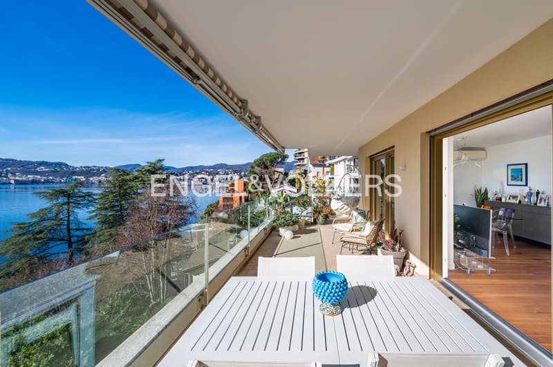 Glass railing, white dining table, white chairs, balcony overlooking water and mountains, plants, wooden floor inside