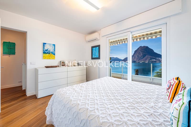 Modern minimalist bedroom with white walls, wooden floor, white bed with knit blanket, large windows with view of water and mountains, white dresser with artwork.