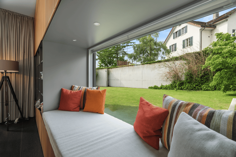 day bed with several pillows, large glass window with window seat, lamp, grass, wall, house outside