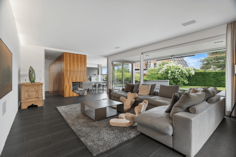 open floor plan, large windows, contemporary design, fireplace, large gray sofa, round coffee table, wooden cabinet, carpet, gray floor tiles