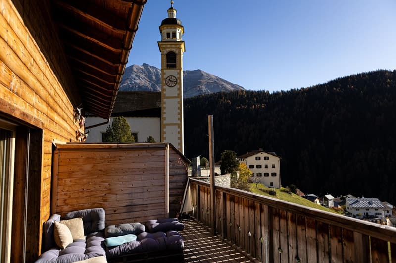 Balcony of a wooden chalet, sofa, clock tower in the background, forest, mountains