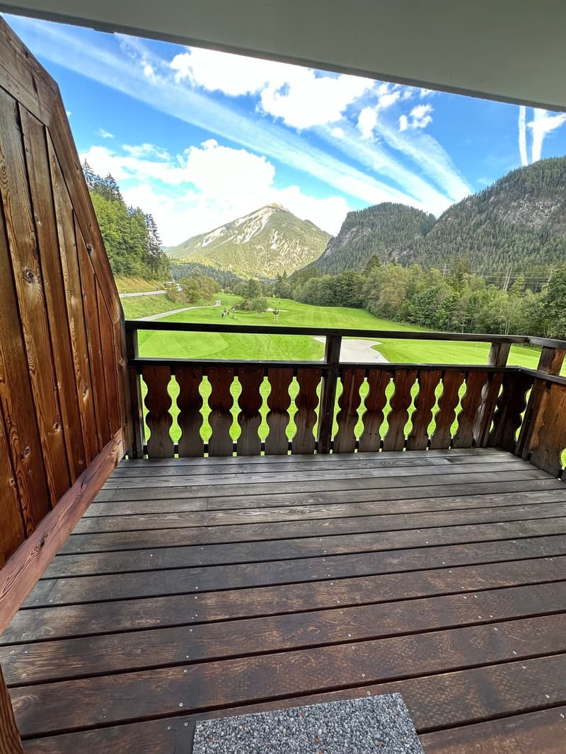 wooden deck, railing, floor rug, mountainous landscape with golf course, lush greenery, blue sky with clouds