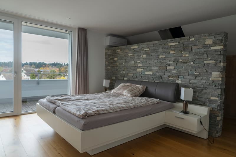 Modern bed, white headboard, wooden floor, double bed, nightstands with lamps, decorative stone wall, sliding glass door leading to balcony, view to outside