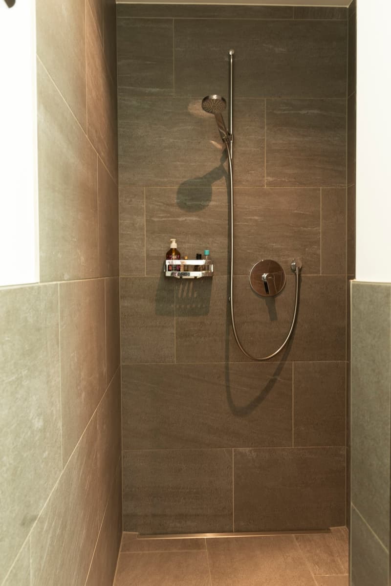 modern shower area with tiled walls and floors, shower head on a flexible arm, small shelf with shampoo