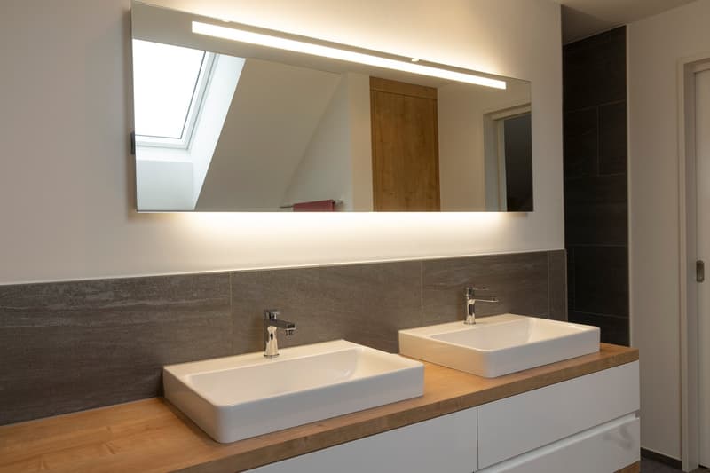 Bathroom with double sinks, mirror with lighting, wooden cabinet, tiled walls, two faucets