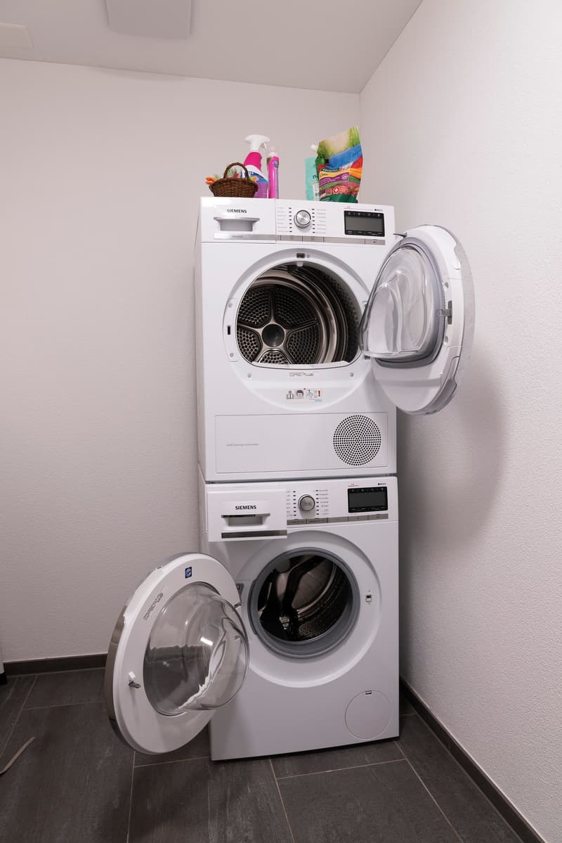 a small room with washing machines stacked on top of each other, and a basket, detergent and cleaning agent on top
