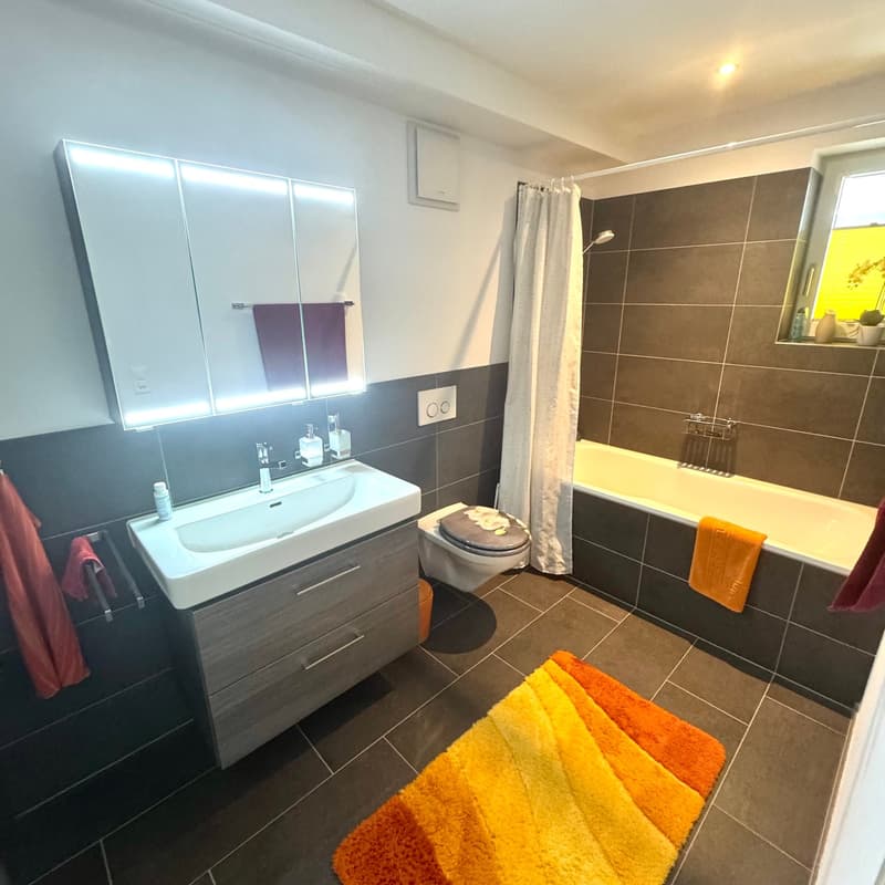 A modern bathroom with a tiled floor, gray and white tiles on the walls, a white vanity with a sink, a mirror, a shower curtain, a white bathtub, and a toilet. There is a white cabinet with drawers, a white towel rack, and a towel on the towel rack. A rug