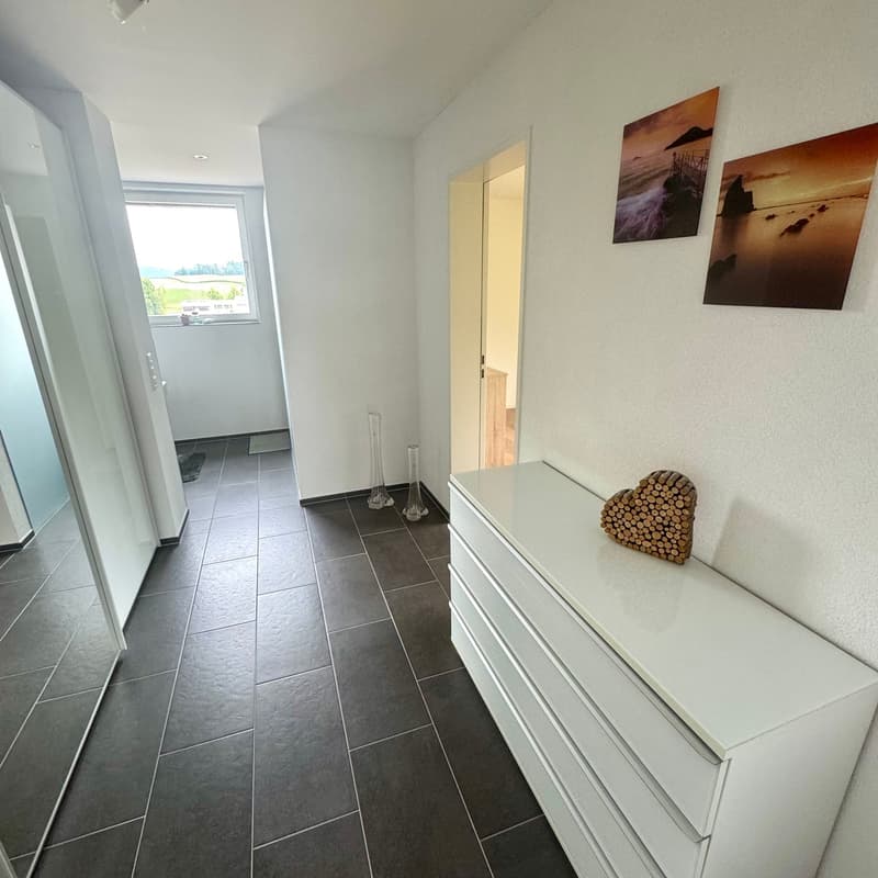 gray tiled floor, white walls, large glass window, full length mirror, two picture frames, two vases, white dresser with decorative log heart on top