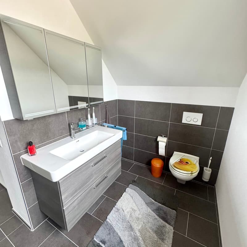 White sink, mirror, dark gray tiles, blue towels, white toilet, silver faucet, gray rug, toiletries on counter.