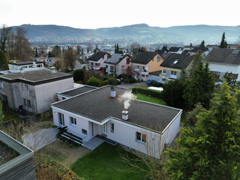 Modern detached house, white exterior, grey gravel roof, two chimneys, surrounded by trees and a green lawn, solar panels on the neighboring house.