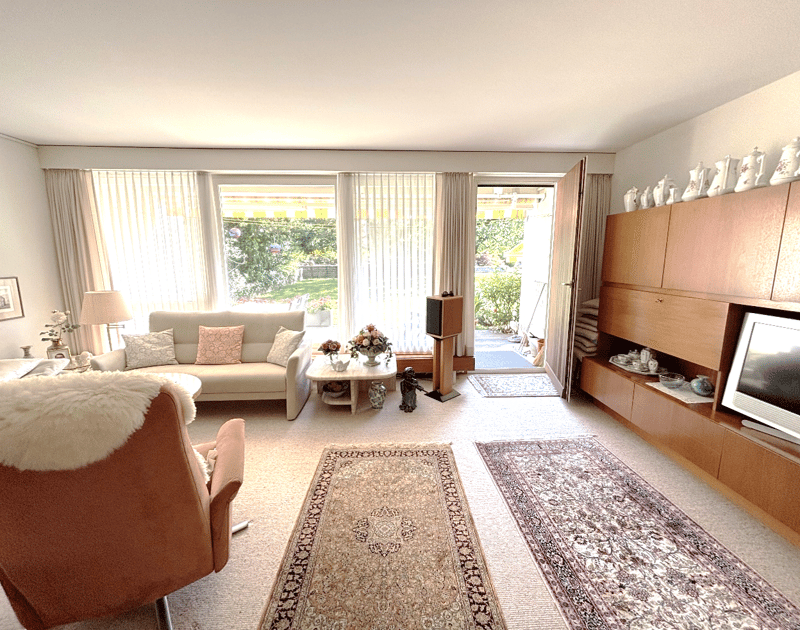 Modern living room, large windows with curtains, white walls, white couch, coffee table, floor, television, shelves