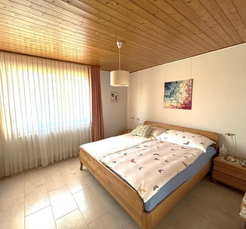 Wooden bed with white sheets and blue blankets, nightstands with lamps, wooden cabinet, picture frames, window with white curtains, wooden ceiling