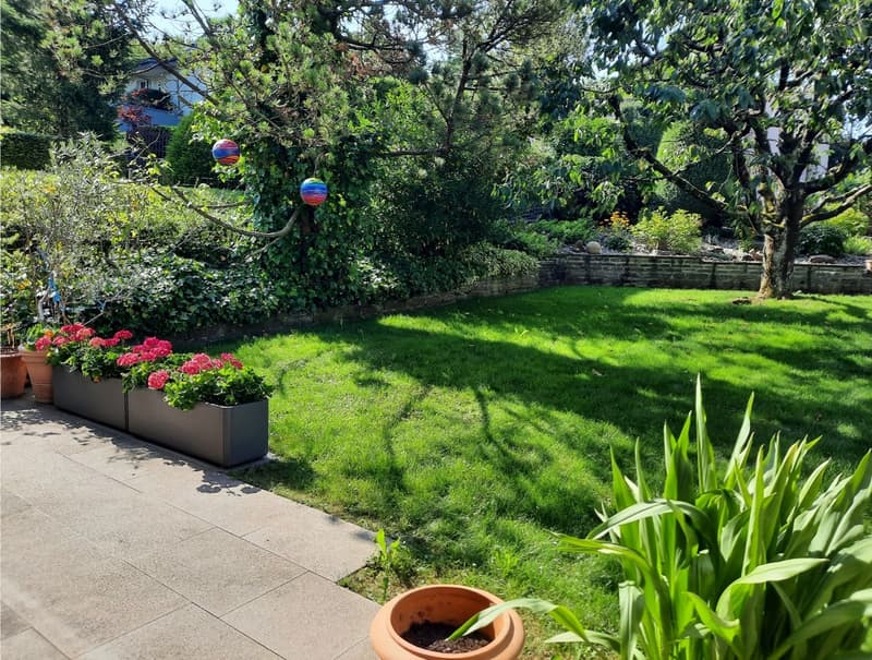 Well-manicured garden with a large lawn, flower beds, trees, and decorative elements like floating balls. The garden is adjacent to a paved pathway.
