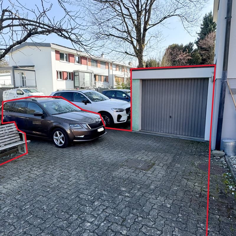 paved parking area with a garage, several cars parked, a bench, a trash can, a building in the background