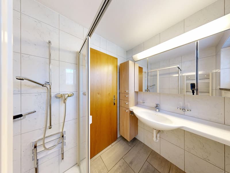 Bathroom with white tiles, wooden door, shower, sink, mirror, wooden cabinet, and towel rack.