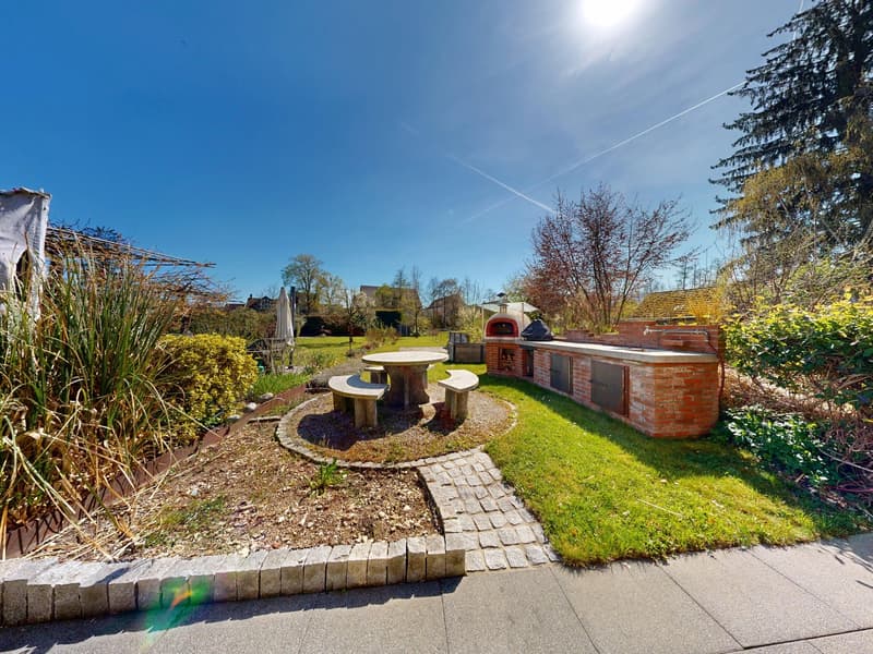 Outdoor dining area with table and benches, brick barbecue, brick wall, green grass, plants, trees, clear blue sky