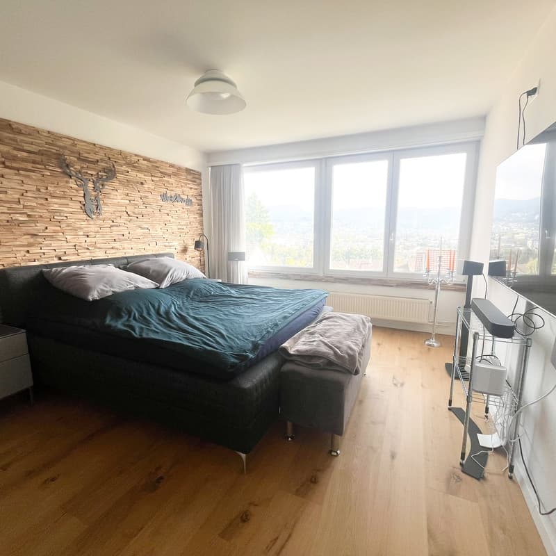 Master bedroom with large double bed, footrest with blanket, wall-mounted TV, windows with curtains, wooden parquet flooring, wooden wall with decorative deer head, ceiling light, night lamps, side table, TV stand, speakers