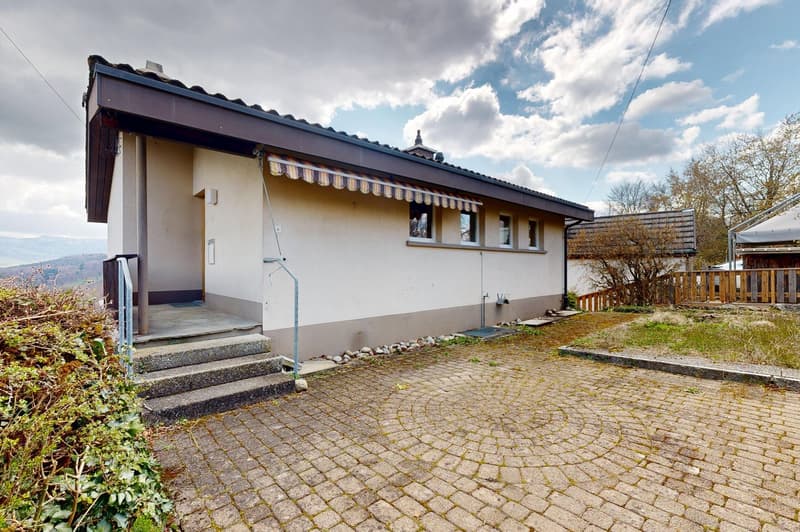 single-story house, white walls, brown roof, brick driveway, awning over the entrance, small garden