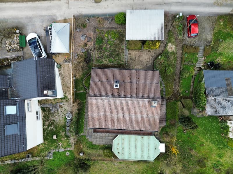 Aerial view of a house with a brown roof, a white car parked in front, a red car parked on the side, a green lawn, and a small shed in the backyard.