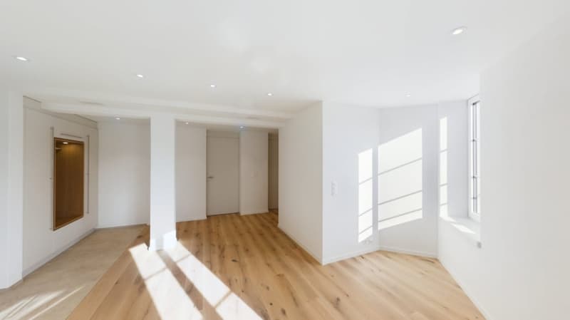 Spacious open-plan living area with hardwood floors, white walls, and large windows allowing plenty of natural light. The room appears to be a combination of a living room and dining area, with a doorway leading to another room or hallway.
