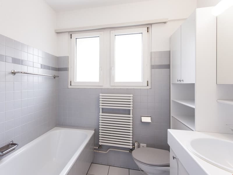 Modern bathroom with bathtub, white tiled walls, white toilet, white sink, white towel warmer, windows, cabinet, and mirror.