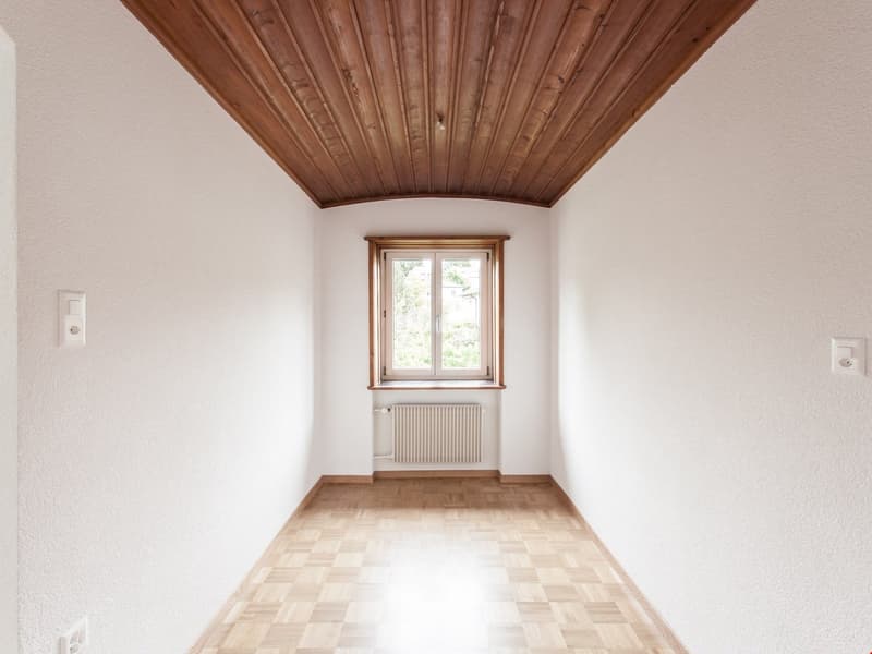 Empty room with wooden ceiling, white walls, wooden parquet floor, window, and white radiator.