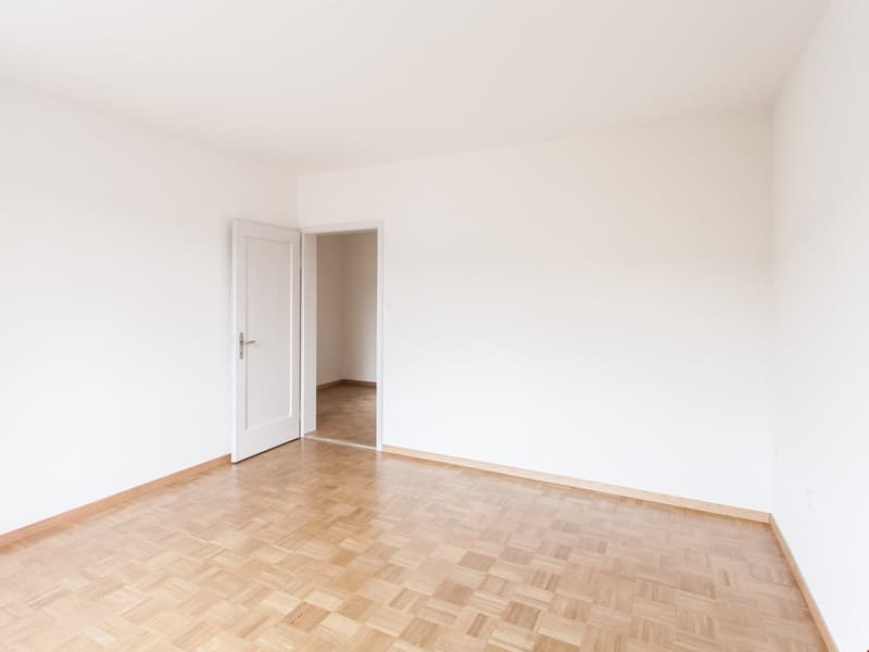 Empty room, wooden parquet floor, white walls, open door leading to another room