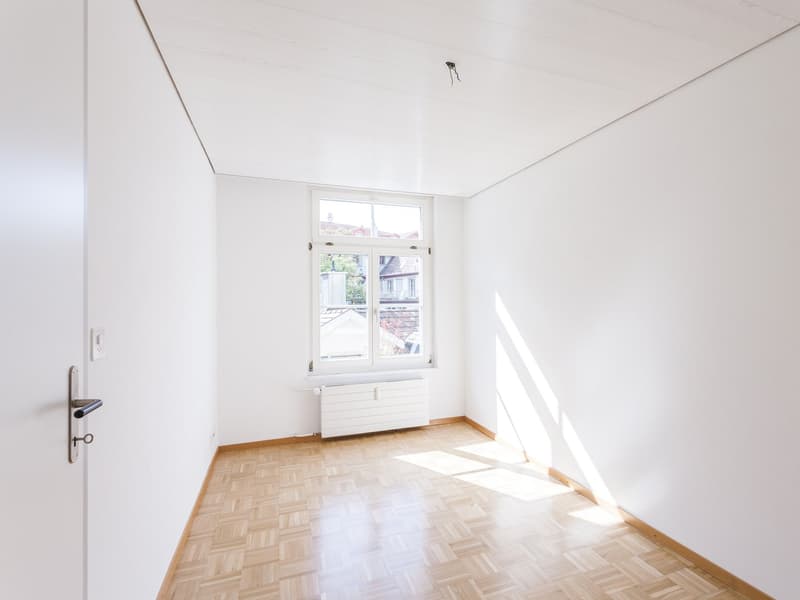 White walls, wooden parquet flooring, double window with wooden frame, a white radiator below the window