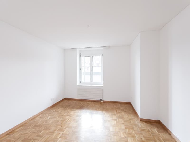 Empty room with white walls, wooden floor, window, and radiator