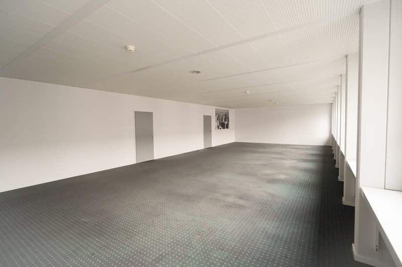 Large open space with white walls, a high ceiling, and a gray tiled floor. The room appears to be empty, with two doors visible on the side walls.