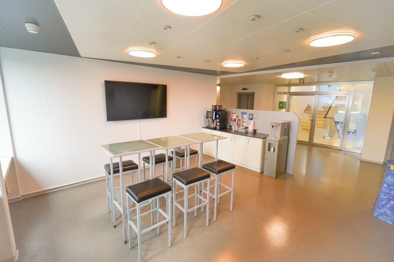 The image shows a modern office or break room space. It has a large TV mounted on the wall, a coffee station with a coffee maker, and a bar-style table with stools. The room has a clean, minimalist design with white walls and recessed lighting. There appe