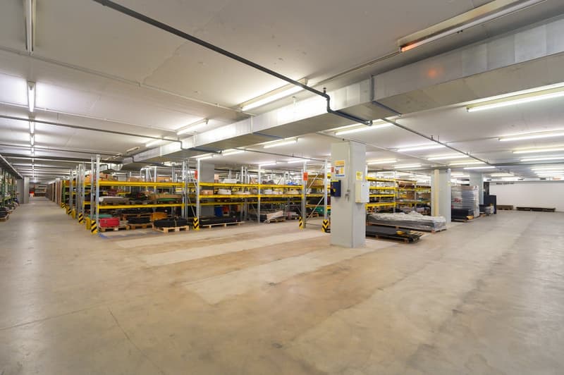 The image shows a large industrial warehouse or storage facility. The space is filled with rows of metal shelving units and storage racks, indicating it is used for storing and organizing various goods and materials. The ceiling is high and the lighting i