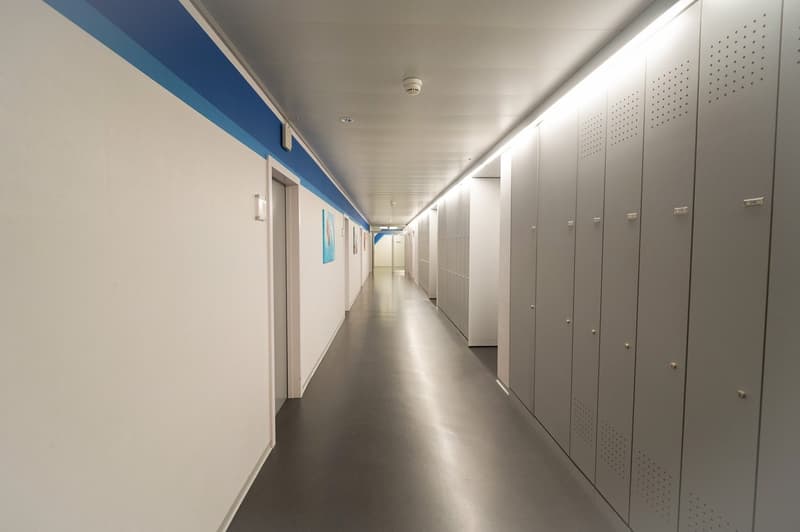 Long, well-lit hallway with white walls, gray lockers or storage units lining the walls, and a blue accent stripe along the ceiling.
