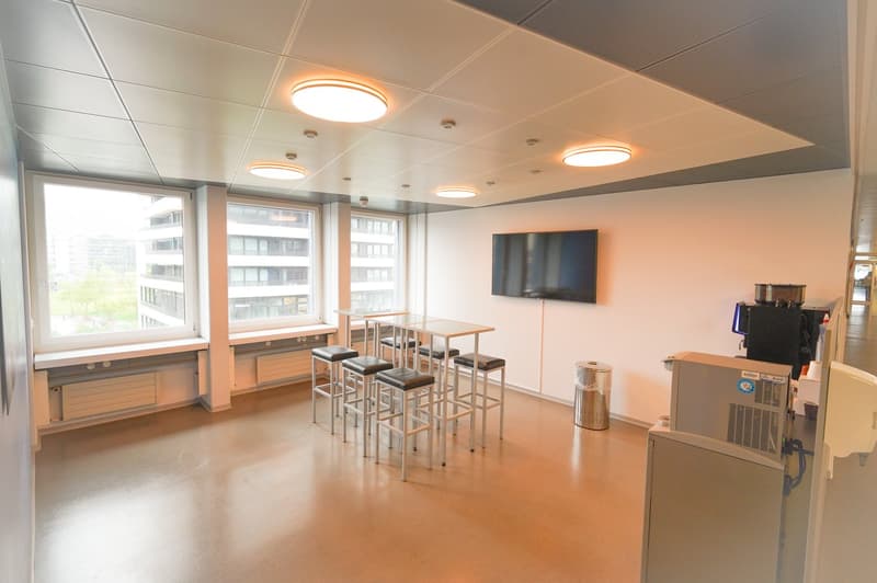 Bright and modern office space with large windows, recessed lighting, bar-style seating, and a TV mounted on the wall. The space appears to be equipped with a dishwasher, likely for employee use.