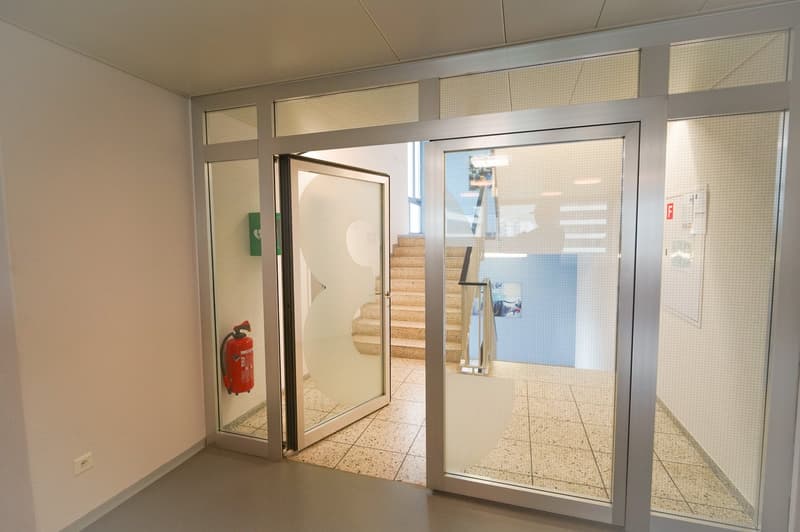 The image shows the interior of a commercial or office space. The space has a glass entrance door leading to a staircase, and there are large windows allowing natural light to enter the space. The walls are painted in a neutral color, and the floor is cov