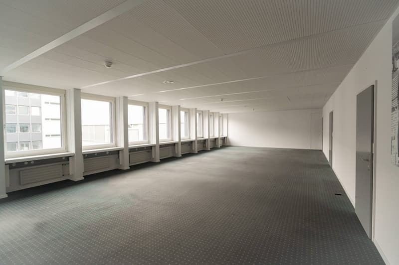 Large open office space with high ceilings, rows of windows, and a gray carpeted floor. The space appears to be empty and ready for use.