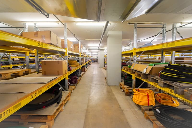 This image shows the interior of a warehouse or storage facility. The space is filled with rows of shelving units and pallets stacked with various boxes and equipment. The shelves are painted in a bright yellow color, and the overall lighting is bright an