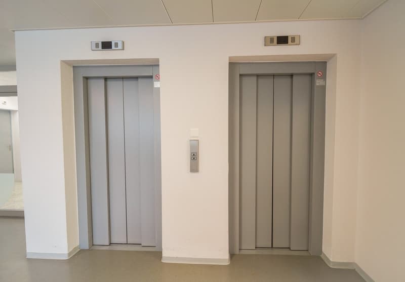 The image shows two elevator doors in a building. The elevators appear to be modern and have a clean, minimalist design with gray panels. There are control panels next to each elevator door, indicating they are functional elevators.