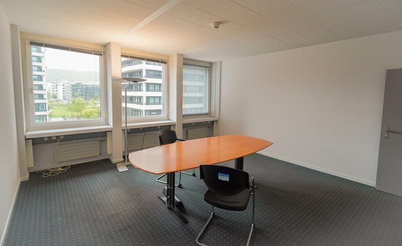 The image shows a modern office space with large windows overlooking a city skyline. The room has a rectangular orange table, black office chairs, and a carpeted floor. The walls are white and the ceiling has recessed lighting fixtures.