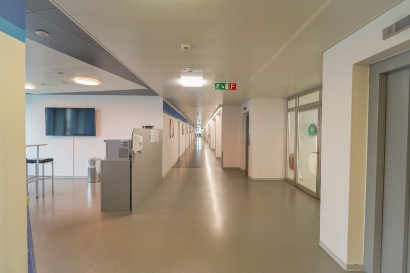 The image shows a long, well-lit hallway in a commercial or institutional building. The hallway has a neutral color scheme with white walls and a gray floor. There are several doors along the hallway, and a television screen mounted on the wall. The hallw