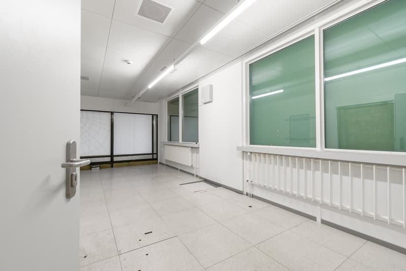 The image shows a large, empty room with white walls, a tiled floor, and fluorescent lighting. There is a projection screen on the wall and glass windows along one side. The room appears to be an office or classroom space.