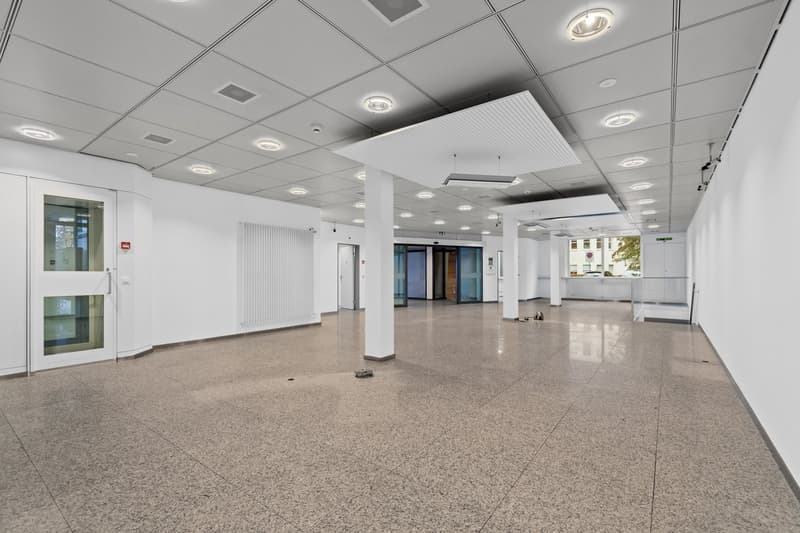The image shows a large, open office space with a high ceiling and a grid of recessed lighting. The floor is made of a light-colored terrazzo material, and the walls are white. There are glass doors and windows visible, as well as a reception area in the